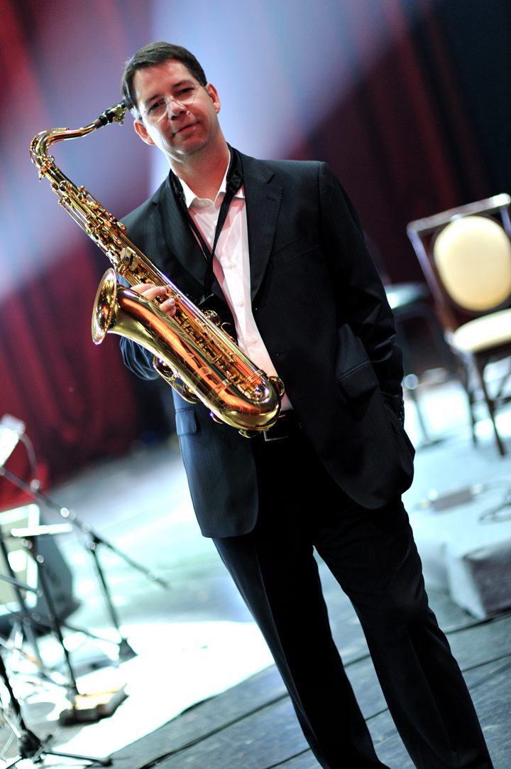 Thomas Hornig on a concert stage in a dark suit, holding a tenor saxophone, with music stands and a chair behind him